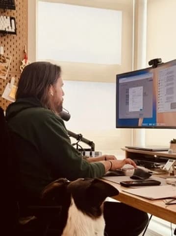 Isaac at his home desk in front of two monitors, with his black and white dog sitting beside him