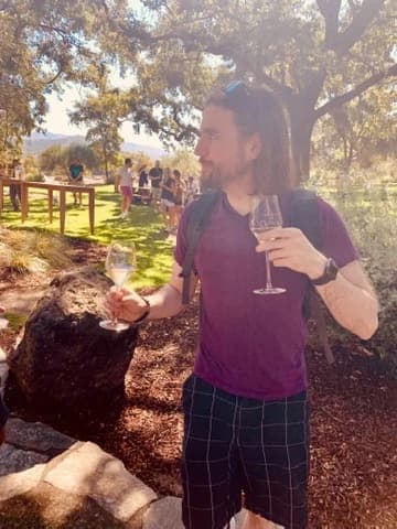 Isaac outdoors under oak trees, holding a glass of white wine in each hand